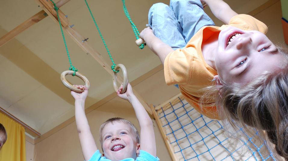 Drei Kinder spielen in einem Raum: Ein Junge hängt an Ringen, während zwei andere lachen und zuschauen.