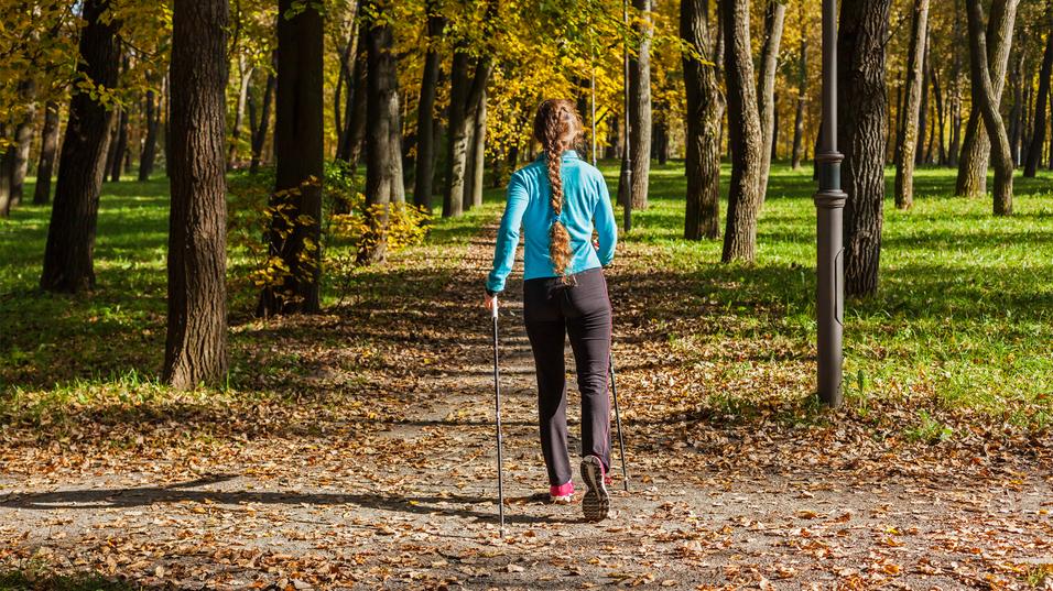 Frau mit langem Zopf geht mit Wanderstöcken einen herbstlichen Weg zwischen Bäumen, umgeben von buntem Laub.