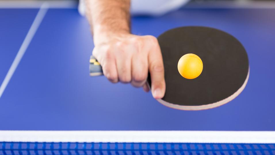 Nahaufnahme einer Hand mit Tischtennisschläger, die einen Tischtennisball über einem blauen Tisch positioniert.